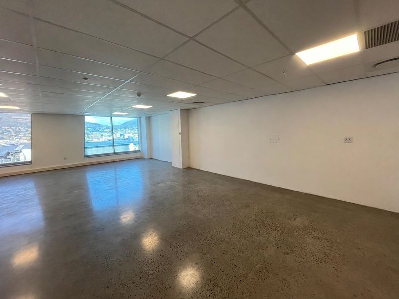 A spacious, empty open-plan office featuring polished concrete floors, suspended ceiling tiles, recessed lighting, and large windows with city views.