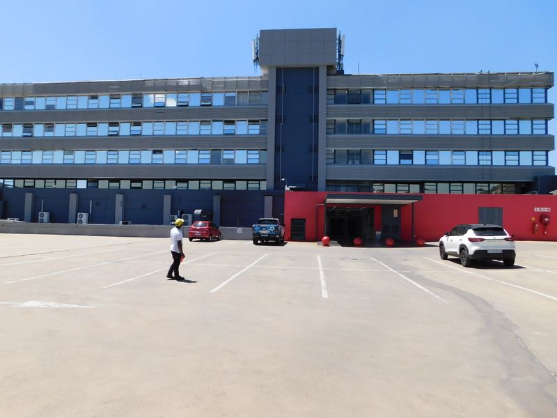 Front exterior view of a multi-story office building featuring a large asphalt parking area and a prominent red-accented entrance.