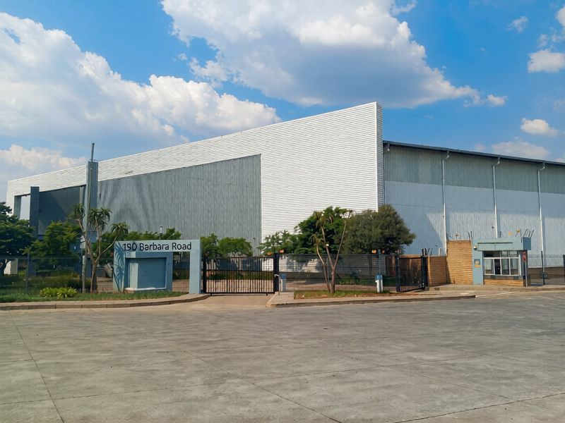 Front view of an industrial property featuring large metal-clad warehouse structures, secure gated entrance, and a dedicated guard house on a paved site.