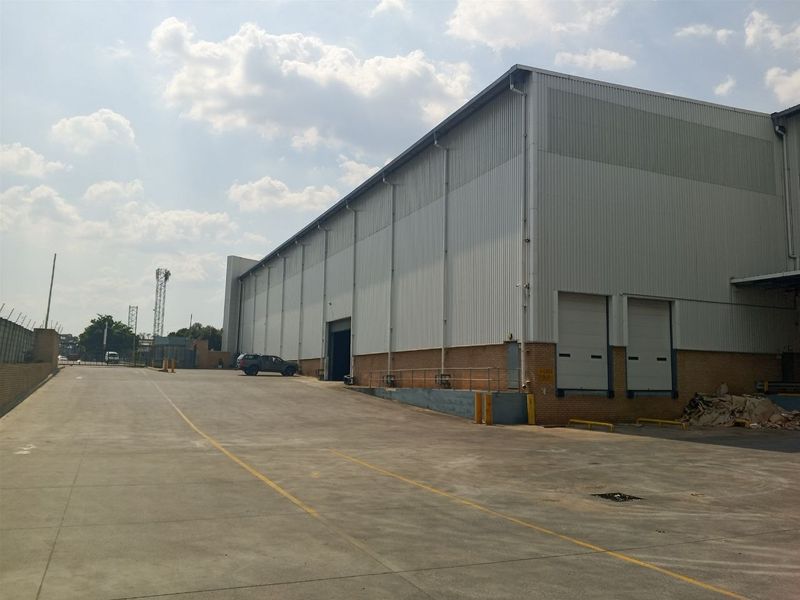 Industrial warehouse exterior featuring corrugated metal cladding, brick base, multiple roller shutter loading doors, and a spacious concrete yard area.