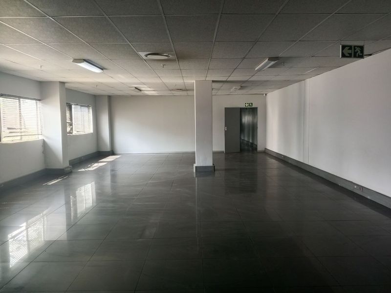A spacious, open-plan industrial office area featuring a suspended ceiling, tiled flooring, and large windows with venetian blinds.