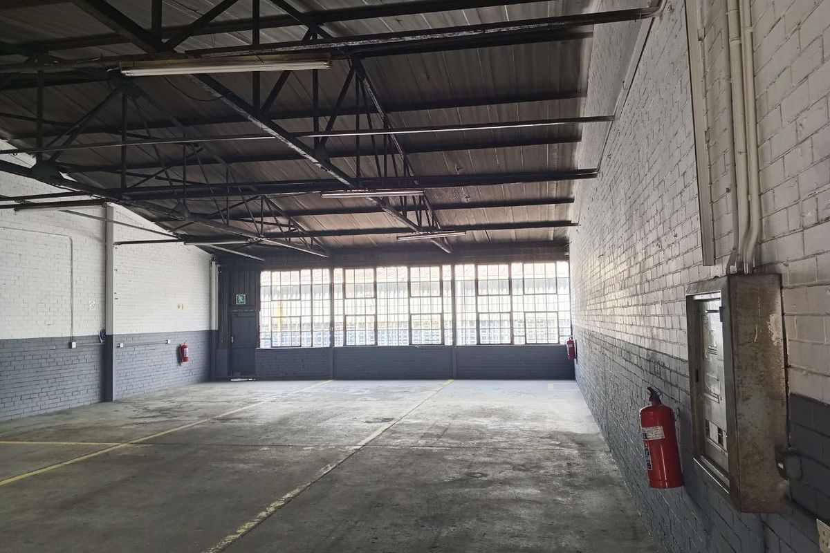 Spacious industrial utility area featuring exposed steel roof trusses, concrete flooring, large metal-framed windows, and painted brick walls, suitable for various commercial or storage uses.