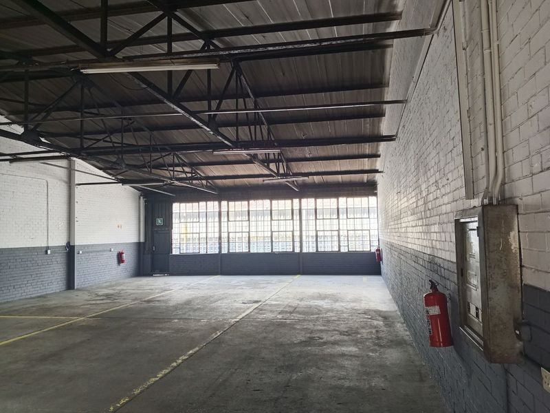 Spacious industrial utility area featuring exposed steel roof trusses, concrete flooring, large metal-framed windows, and painted brick walls, suitable for various commercial or storage uses.