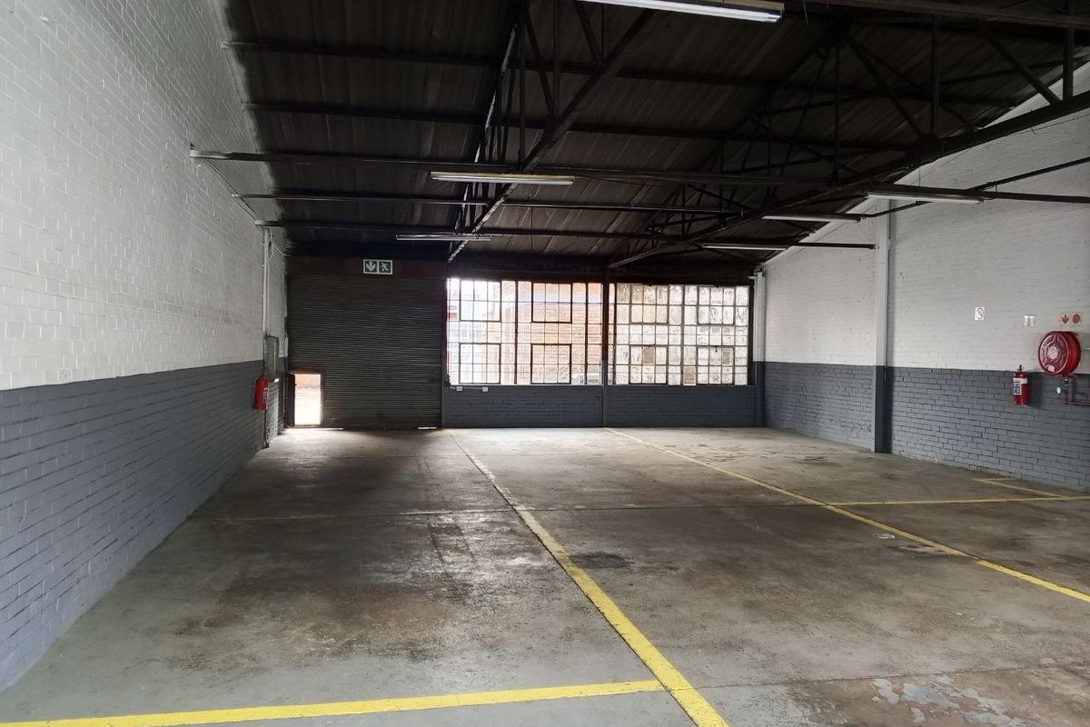 Spacious industrial warehouse unit featuring concrete flooring, exposed steel roof trusses, large steel-framed windows, and a secure roller shutter door.