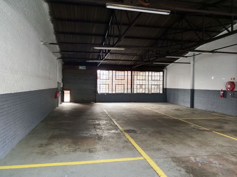Spacious industrial warehouse unit featuring concrete flooring, exposed steel roof trusses, large steel-framed windows, and a secure roller shutter door.
