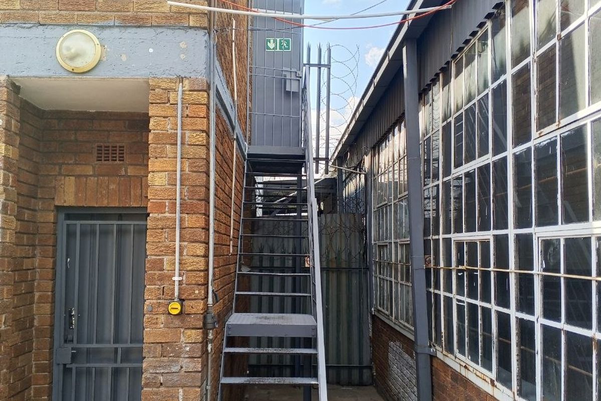 Exterior view of an industrial property featuring a metal fire escape staircase, security fencing, and large steel-framed windows along the side elevation.