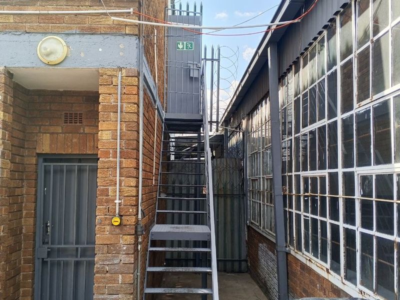 Exterior view of an industrial property featuring a metal fire escape staircase, security fencing, and large steel-framed windows along the side elevation.