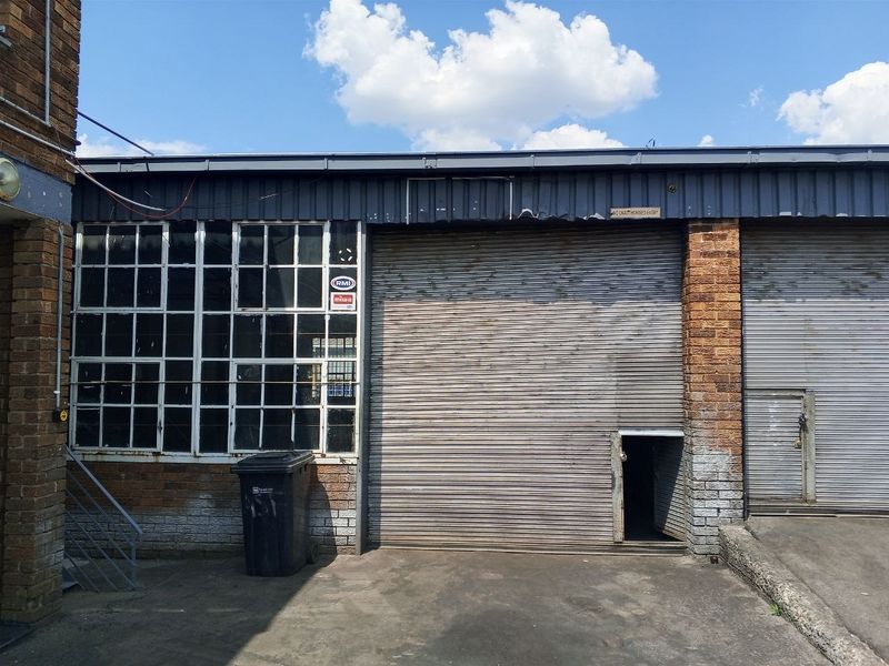 Industrial exterior featuring a roller shutter door, steel-framed windows, and brick facade, showing signs of age and wear.