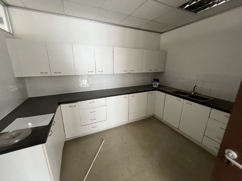 A functional office kitchenette featuring white cabinetry, dark laminate countertops, a double stainless steel sink, and tiled splashbacks in a clean, empty space.