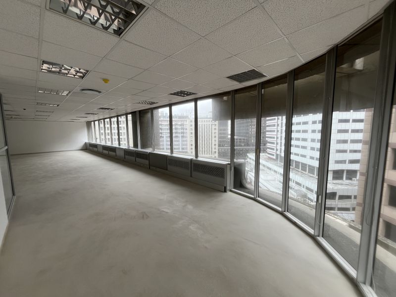 A spacious, empty open-plan office area featuring a suspended ceiling, recessed lighting, and extensive floor-to-ceiling windows offering an urban city view.