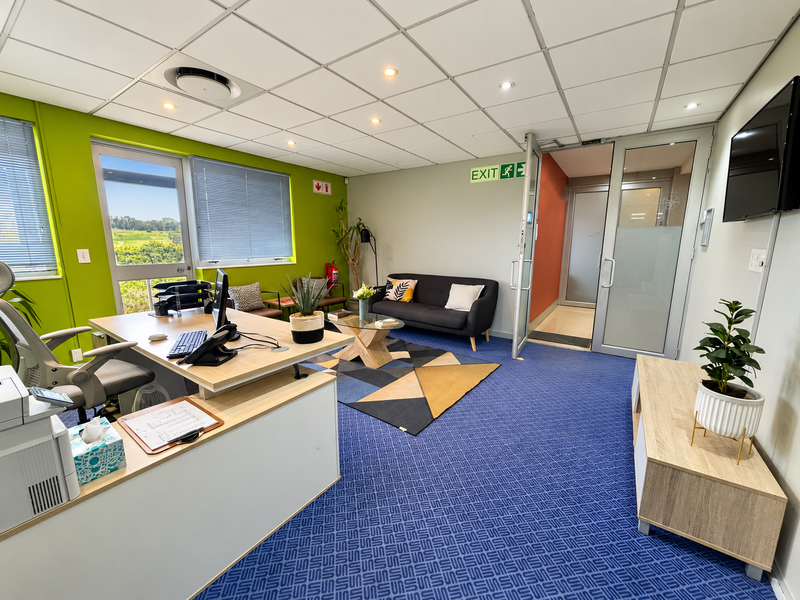 A well-lit professional office space featuring a suspended ceiling, large windows with natural light, blue carpeting, and a glass-panelled entrance door.