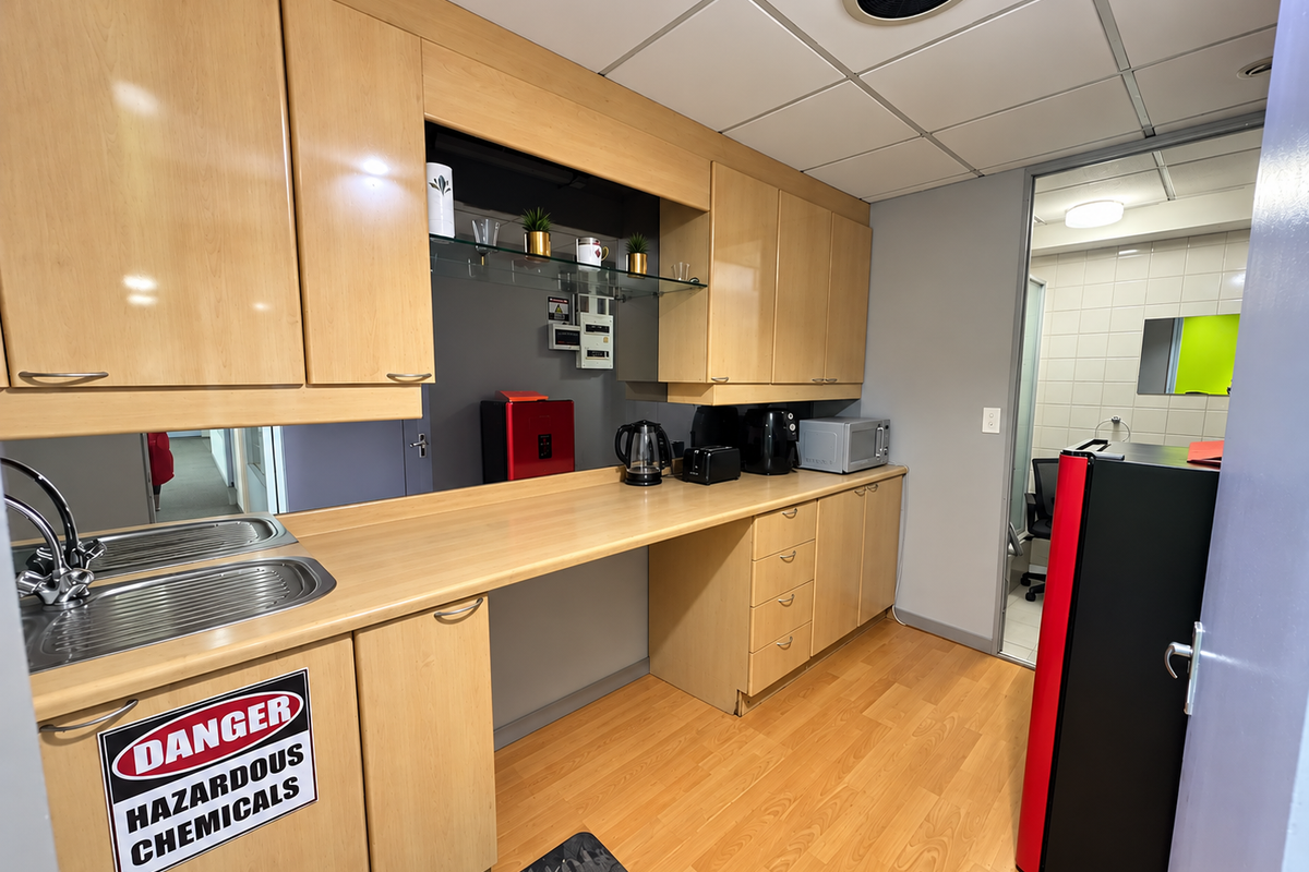 A functional office kitchenette featuring light wood-tone cabinetry, laminate countertops, a stainless steel sink, and a drop ceiling with integrated ventilation.