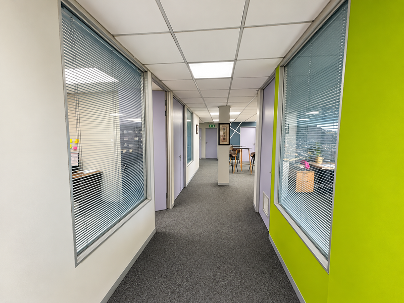 A bright office hallway featuring carpeted flooring, suspended ceilings with recessed lighting, and glass partitions providing visibility into adjacent office spaces.