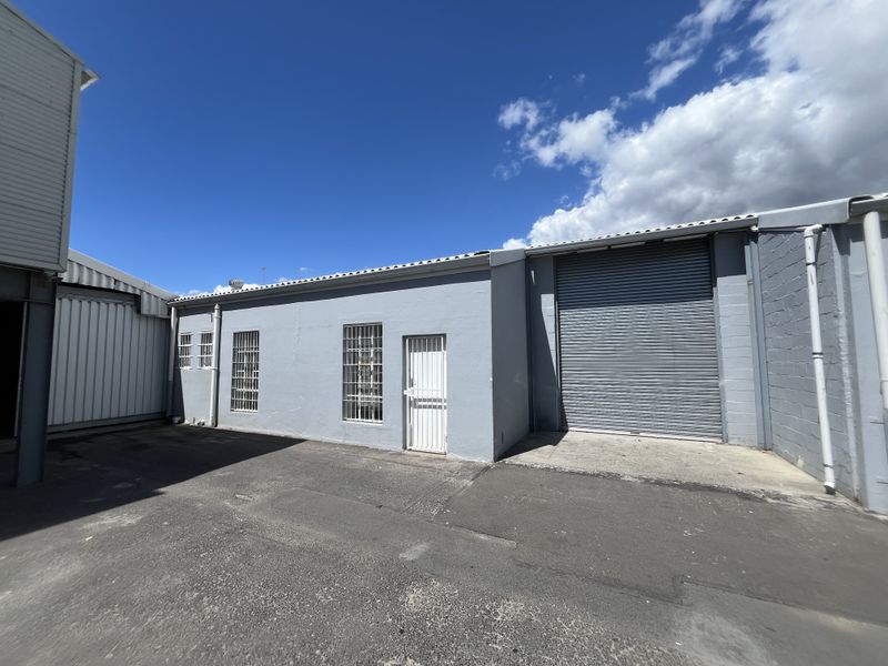 Rear exterior view of an industrial warehouse featuring a large roller shutter door, security-barred windows, and an asphalt yard area.