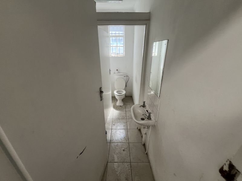 A compact industrial-style bathroom featuring a wall-mounted basin, toilet, and tiled flooring, requiring cosmetic updates to improve its overall condition.