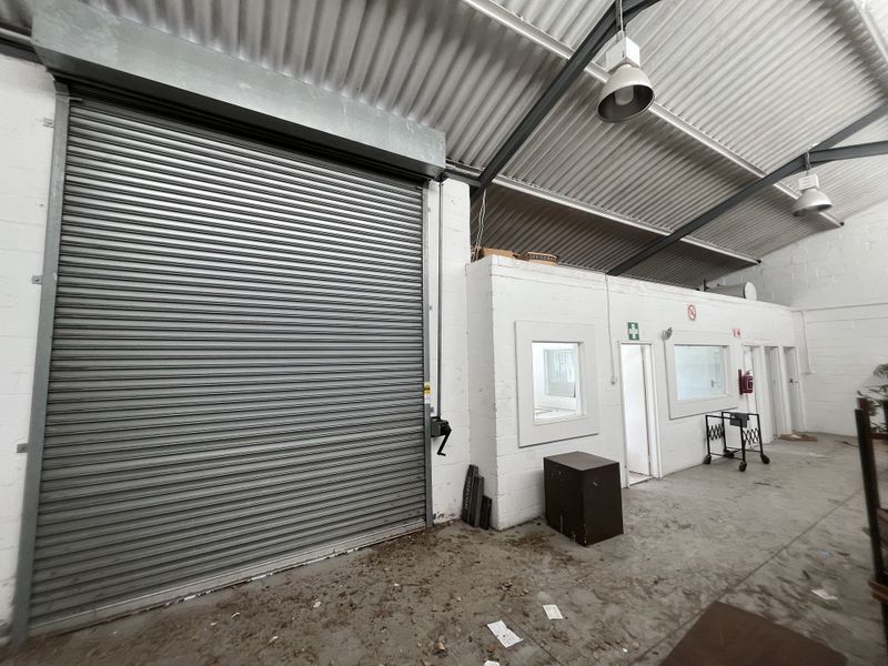 Spacious industrial utility area featuring a large roller shutter door, high ceilings with exposed steel trusses, and integrated office partitioning.