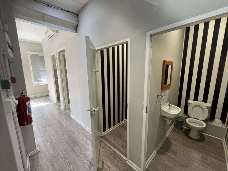 A functional office hallway featuring laminate flooring, a wall-mounted air conditioning unit, and access to a small bathroom facility.