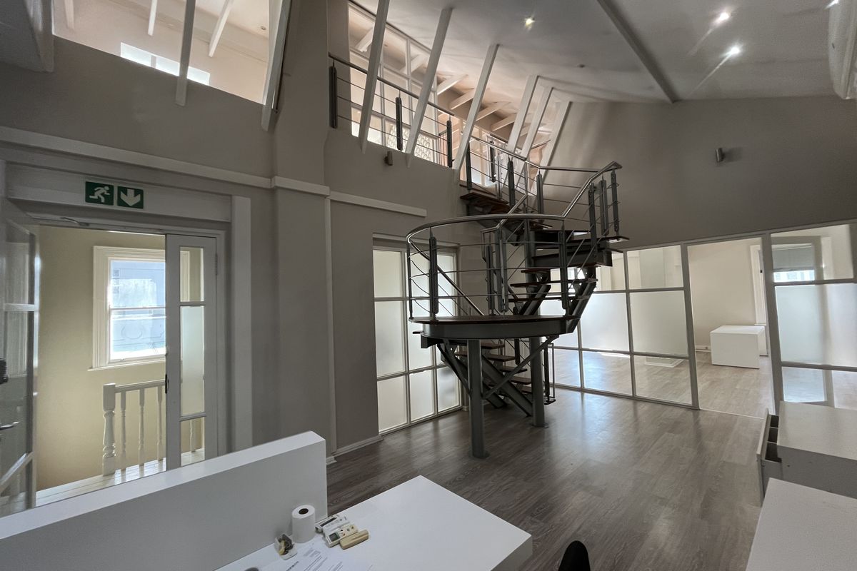 Spacious open-plan office featuring a striking spiral staircase leading to a mezzanine level, glass partitions, and high ceilings with exposed roof trusses.