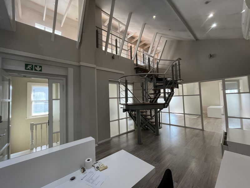 Spacious open-plan office featuring a striking spiral staircase leading to a mezzanine level, glass partitions, and high ceilings with exposed roof trusses.