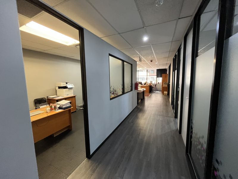 A professional office hallway featuring modern laminate flooring, suspended ceilings with recessed lighting, and glass partition walls leading into private office spaces.