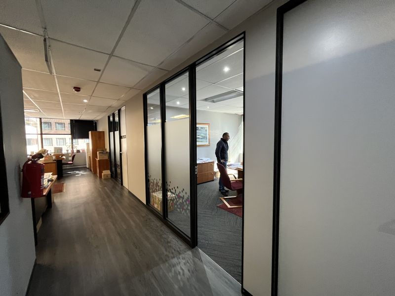 Office corridor featuring glass partition walls, suspended ceilings, and a mix of laminate and carpet flooring in a professional workspace setting.