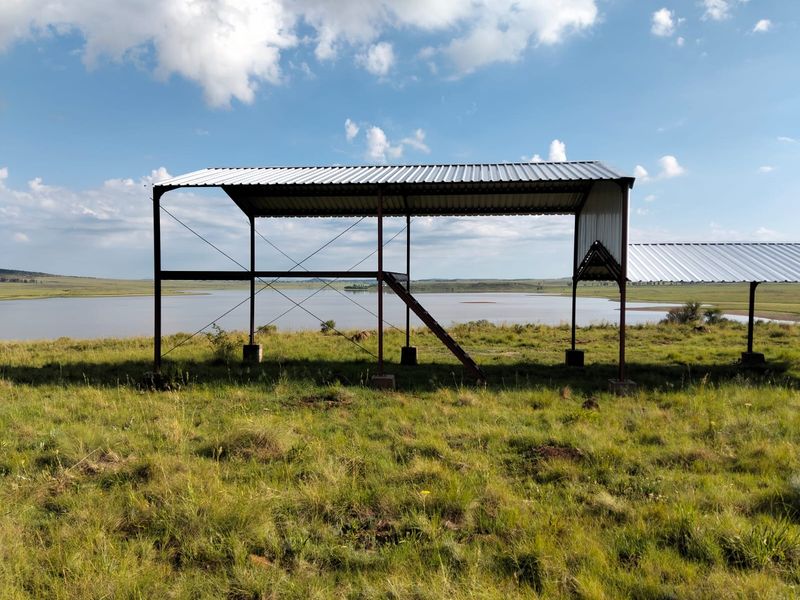 An open-sided metal frame structure situated on a grassy field overlooking a large dam or lake.