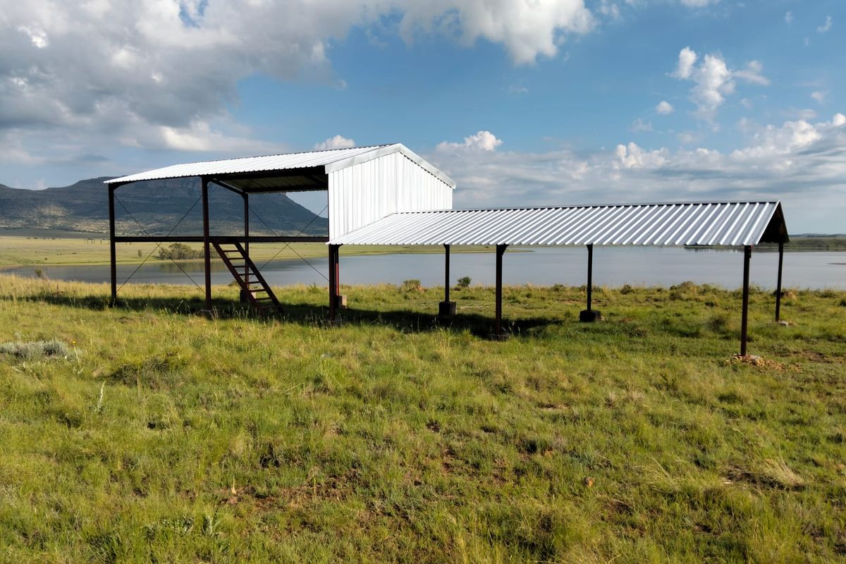 An unfinished metal structure with a raised platform and staircase, situated in an open field overlooking a large dam and mountain landscape.