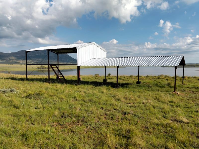 An unfinished metal structure with a raised platform and staircase, situated in an open field overlooking a large dam and mountain landscape.