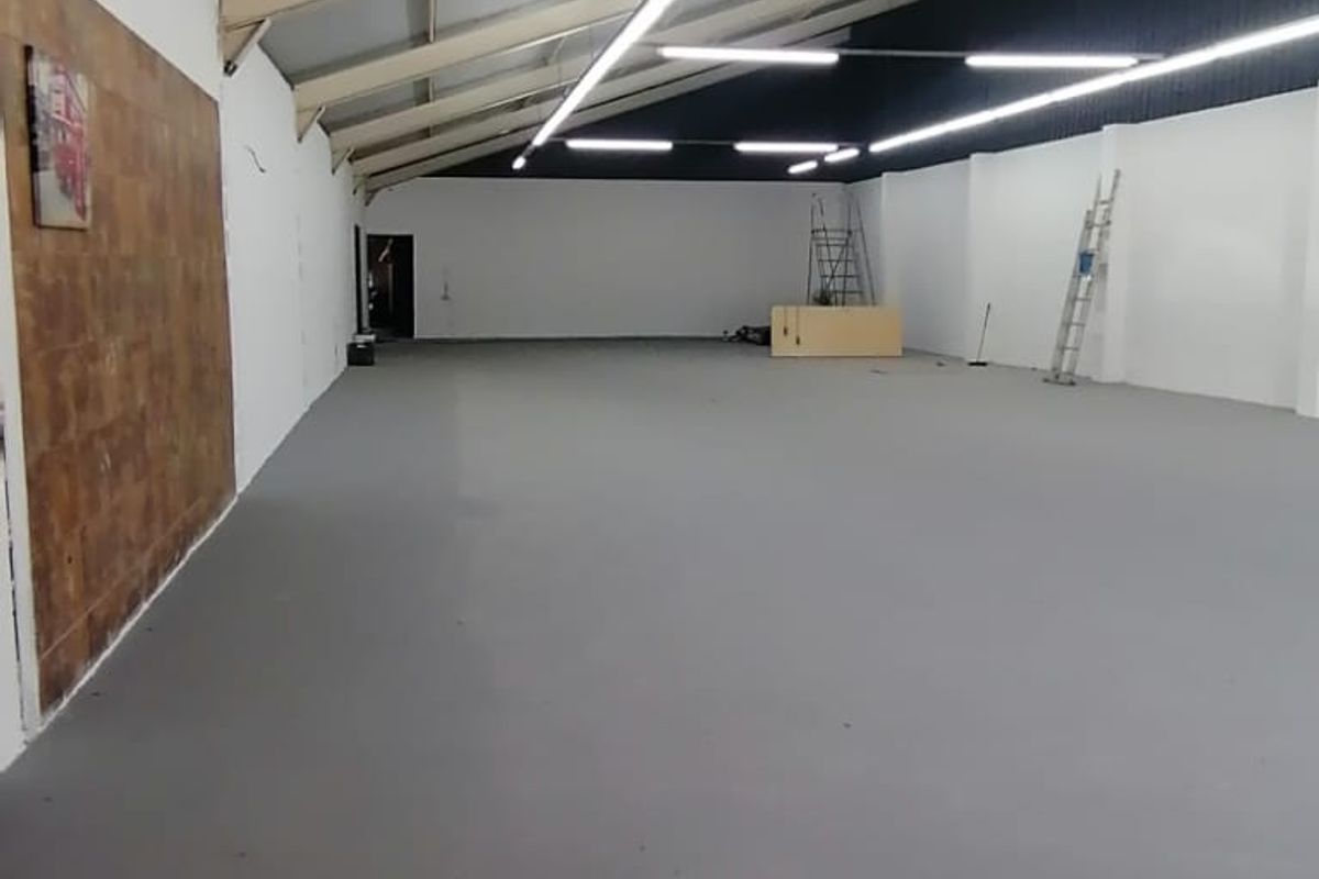 Spacious open-plan retail or commercial area featuring high ceilings with exposed steel trusses, modern strip lighting, and a neutral painted concrete floor.