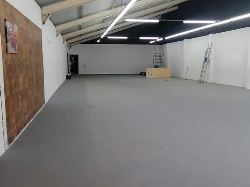 Spacious open-plan retail or commercial area featuring high ceilings with exposed steel trusses, modern strip lighting, and a neutral painted concrete floor.