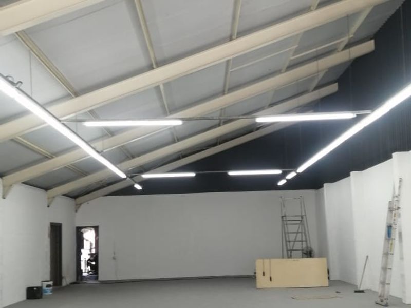 A spacious, open-plan utility or warehouse area featuring high ceilings with exposed steel trusses, bright fluorescent lighting, and clean concrete flooring.