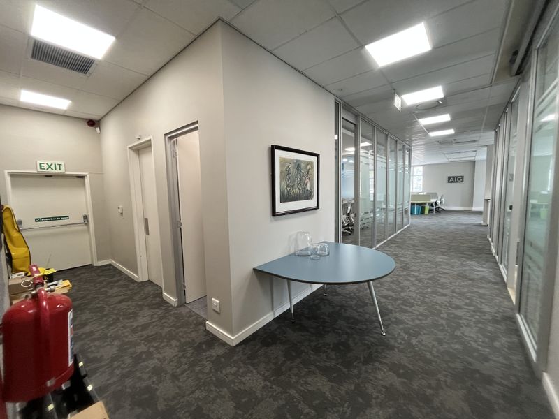 Modern office corridor featuring glass partition walls, suspended ceilings with recessed lighting, and professional carpet flooring.