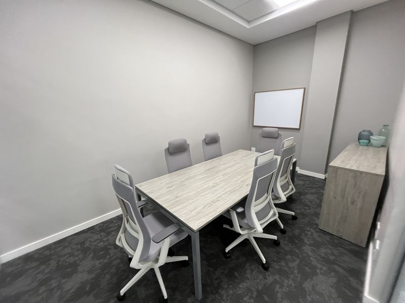 A modern, well-maintained office meeting room featuring neutral grey walls, recessed ceiling lighting, a wall-mounted whiteboard, and dark patterned carpeting.