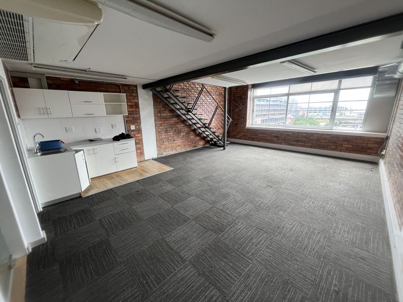 Open-plan office space featuring exposed brick walls, industrial-style steel staircase, kitchenette, and large windows providing ample natural light.