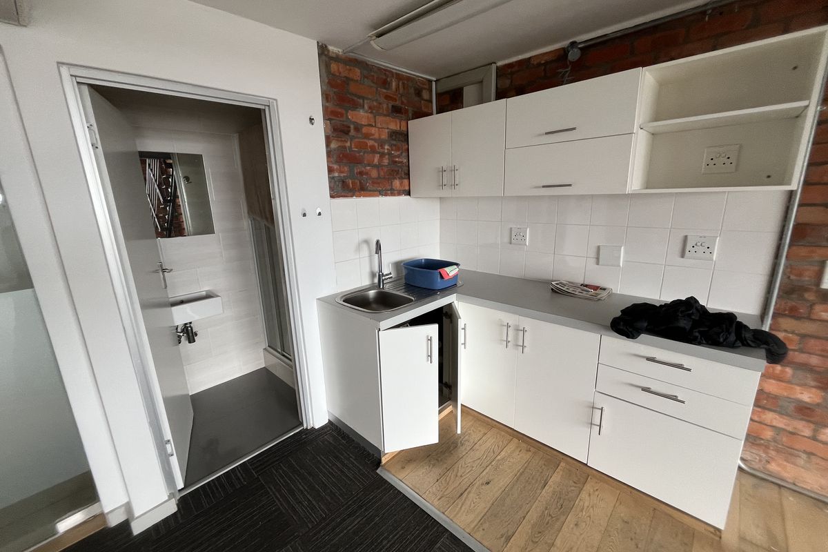 A compact office kitchenette featuring white cabinetry, a stainless steel sink, tiled backsplash, and exposed brick accents, adjacent to a small private restroom.