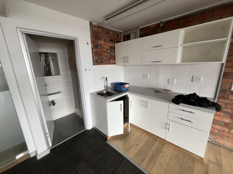 A compact office kitchenette featuring white cabinetry, a stainless steel sink, tiled backsplash, and exposed brick accents, adjacent to a small private restroom.