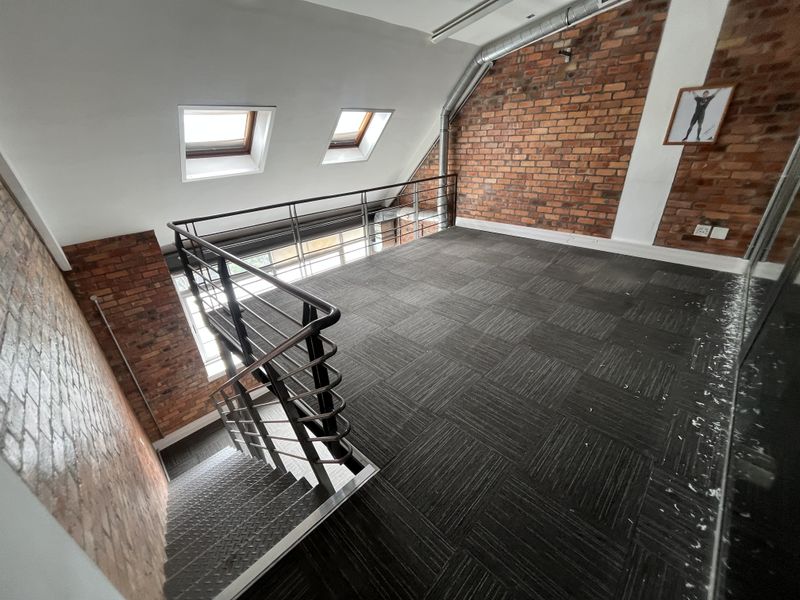 An open-plan office mezzanine level featuring exposed brick walls, skylights, and a metal staircase with a modern industrial aesthetic.