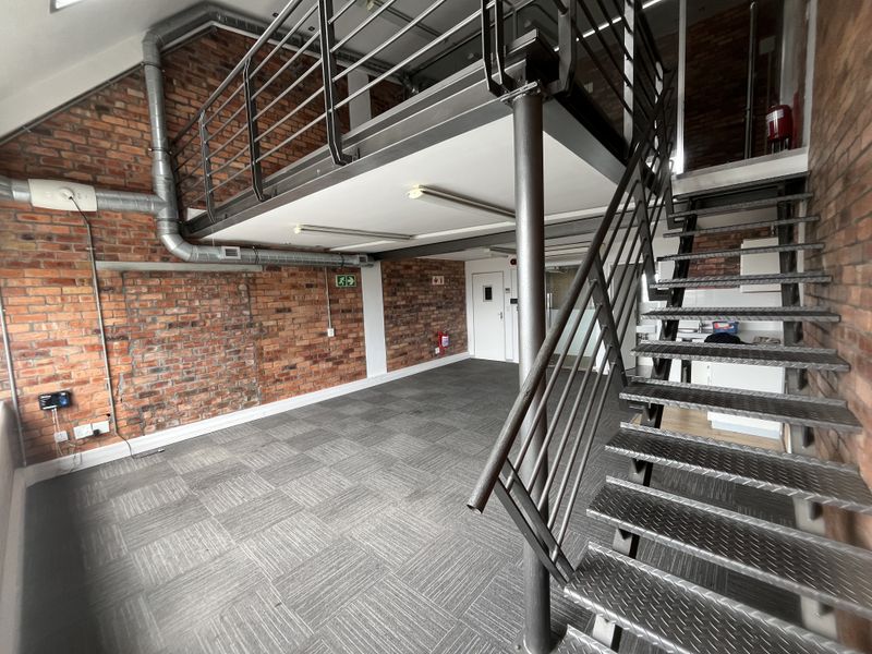 Modern industrial-style office space featuring exposed brick walls, a mezzanine level with a metal staircase, and practical carpet tile flooring.