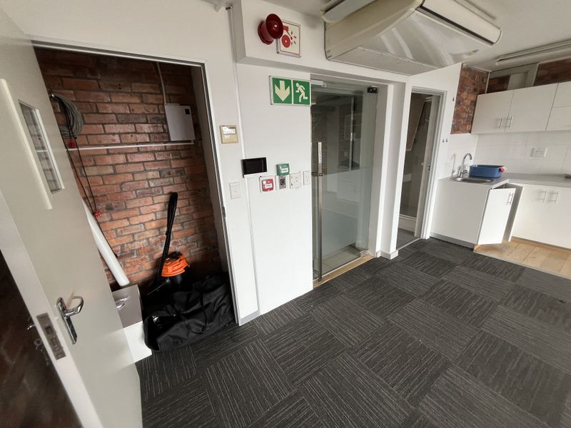 An office space featuring carpet tiles, an exposed brick wall, a small kitchenette with built-in cabinetry, and a glass-paneled door.
