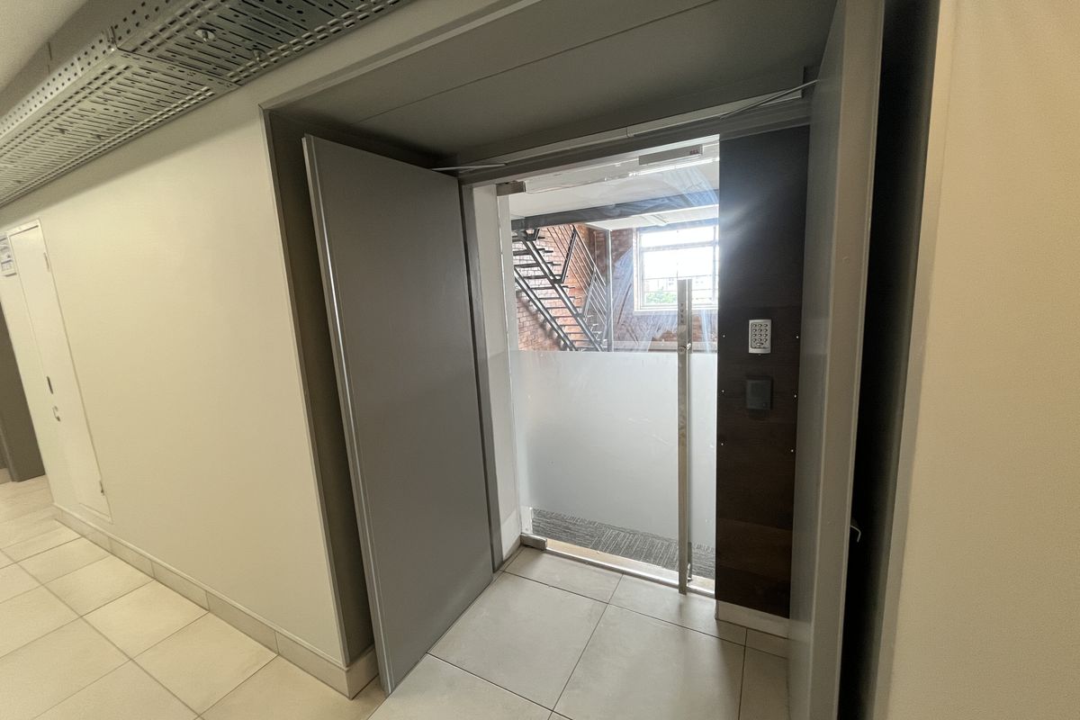 A modern office hallway featuring tiled flooring, exposed cable management, and a secure glass door with keypad access leading to an external staircase.