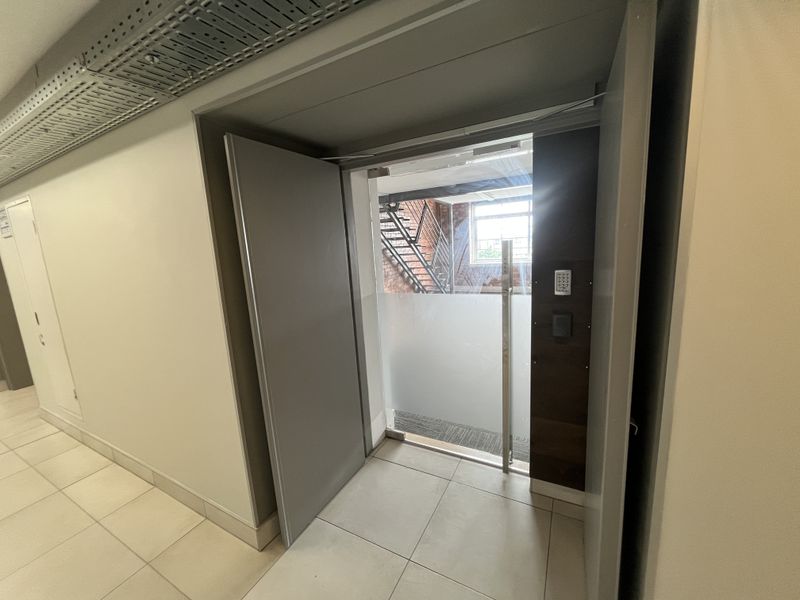 A modern office hallway featuring tiled flooring, exposed cable management, and a secure glass door with keypad access leading to an external staircase.