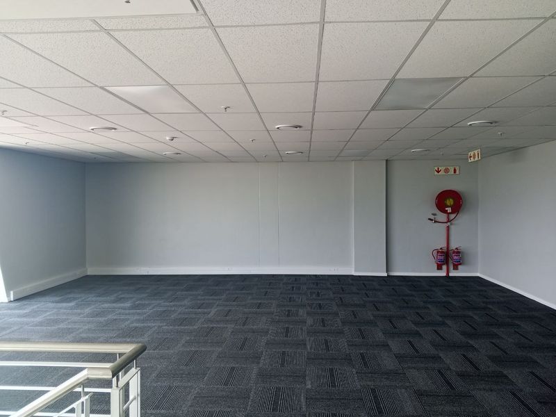 A spacious, empty office area featuring a suspended ceiling, recessed lighting, and dark carpet tiles, equipped with essential fire safety equipment.