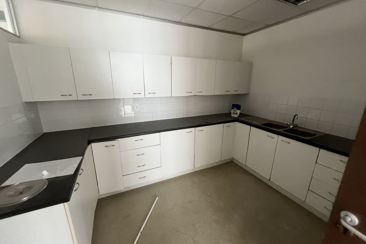 A functional office kitchenette featuring white cabinetry, dark laminate countertops, a double stainless steel sink, and tiled splashbacks in a clean, empty space.