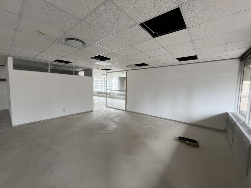 An empty, open-plan office space featuring glass partitions, large windows, and a suspended ceiling with several missing tiles.