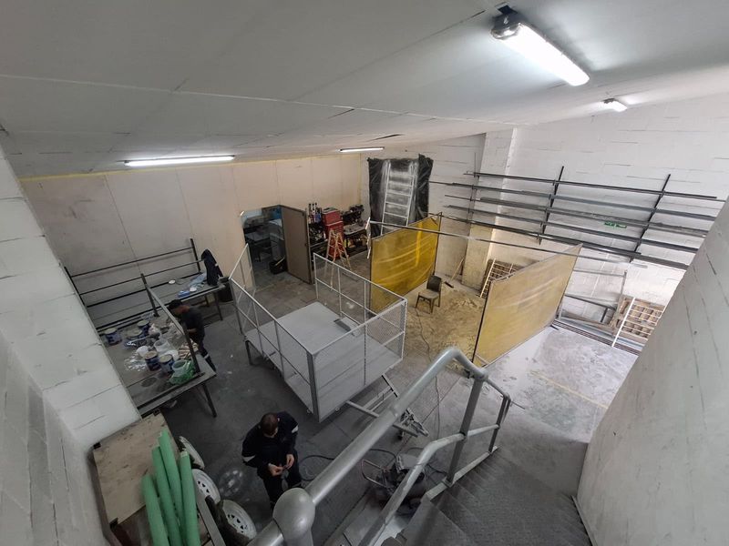 Industrial workshop space featuring concrete flooring, metal wall shelving, and overhead fluorescent lighting, suitable for manufacturing or storage operations.