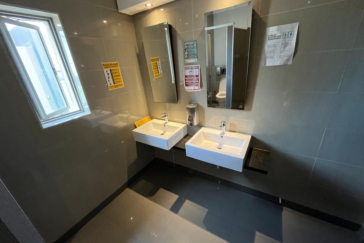 Modern office bathroom featuring dual wall-mounted ceramic basins, sleek chrome faucets, and large rectangular mirrors with tiled walls and flooring.