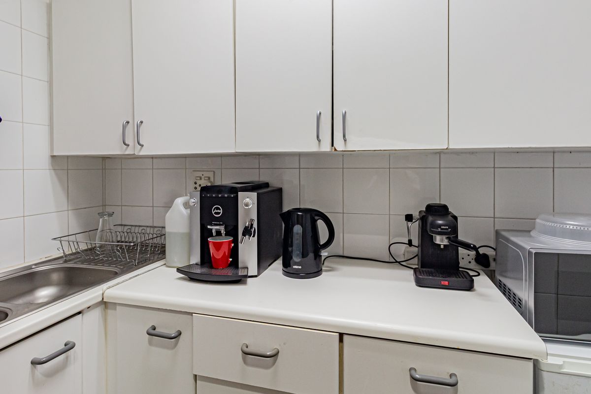 A functional office kitchenette featuring white cabinetry, a stainless steel sink, and tiled splashbacks, providing essential utility for a professional workspace.