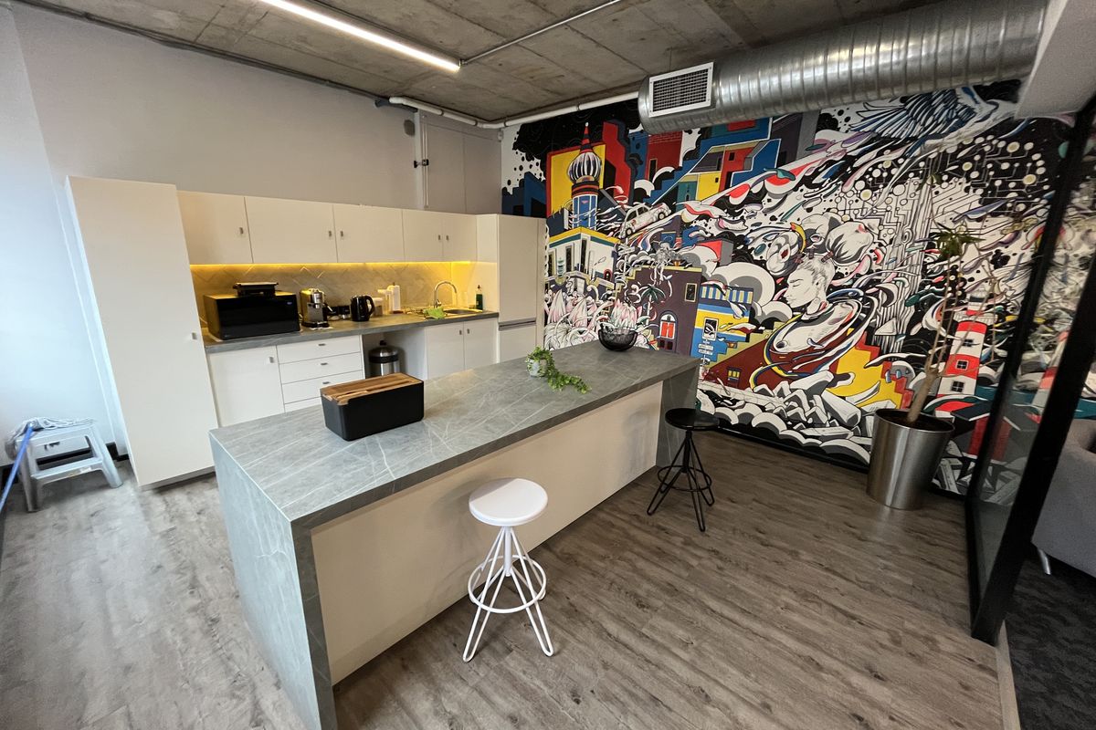 Modern office kitchenette featuring a stone-topped island, sleek white cabinetry, exposed concrete ceiling with industrial ducting, and a vibrant feature wall mural.