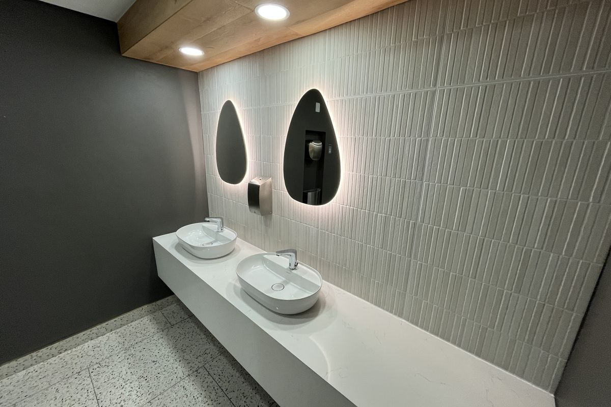 Modern office restroom featuring a floating vanity with dual vessel sinks, backlit teardrop mirrors, and textured wall tiling.