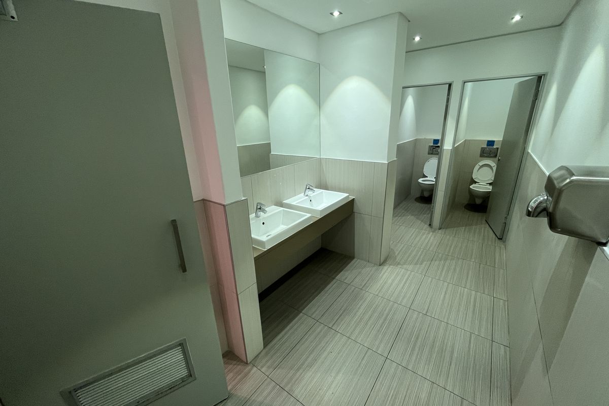 Modern office restroom featuring dual vanity sinks, large wall-mounted mirrors, and separate toilet cubicles with tiled flooring and recessed lighting.